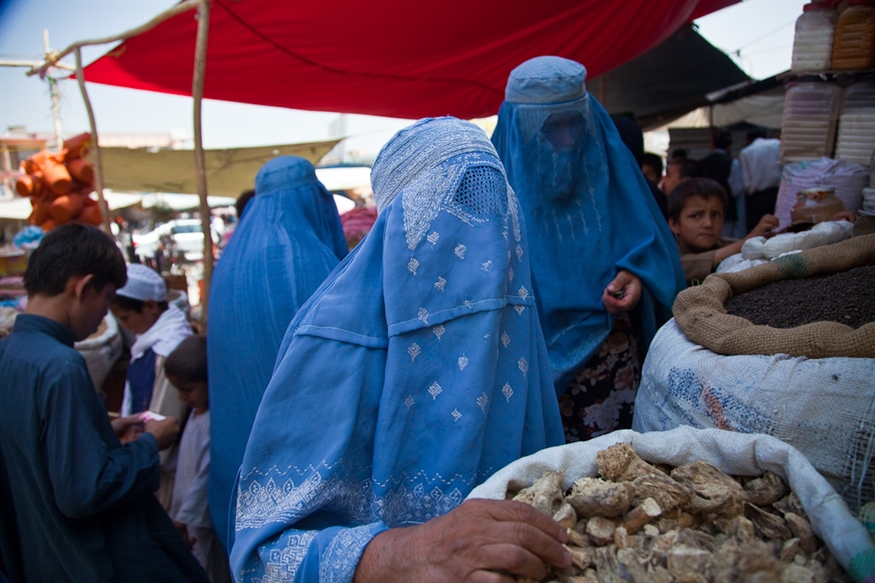 [ai] Two women wearing blue burqas in a market, with one woman reaching towards bags of goods. Children and other shoppers are visible in the background under colorful market awnings.